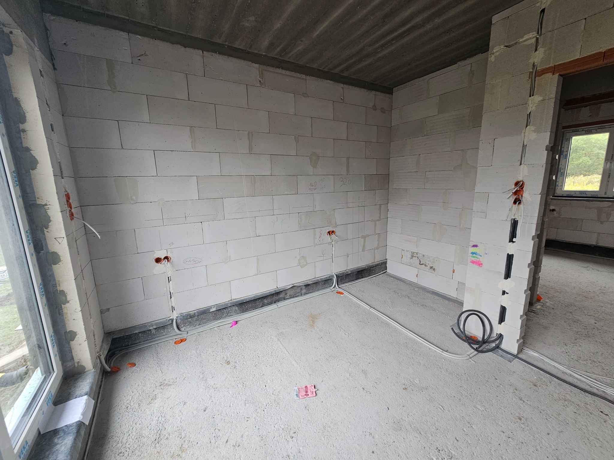 Wnętrze w stanie surowym z widoczną instalacją elektryczną w peszlach i puszkach, ściany z białego betonu komórkowego, okno z widokiem na zieleń na zewnątrz.