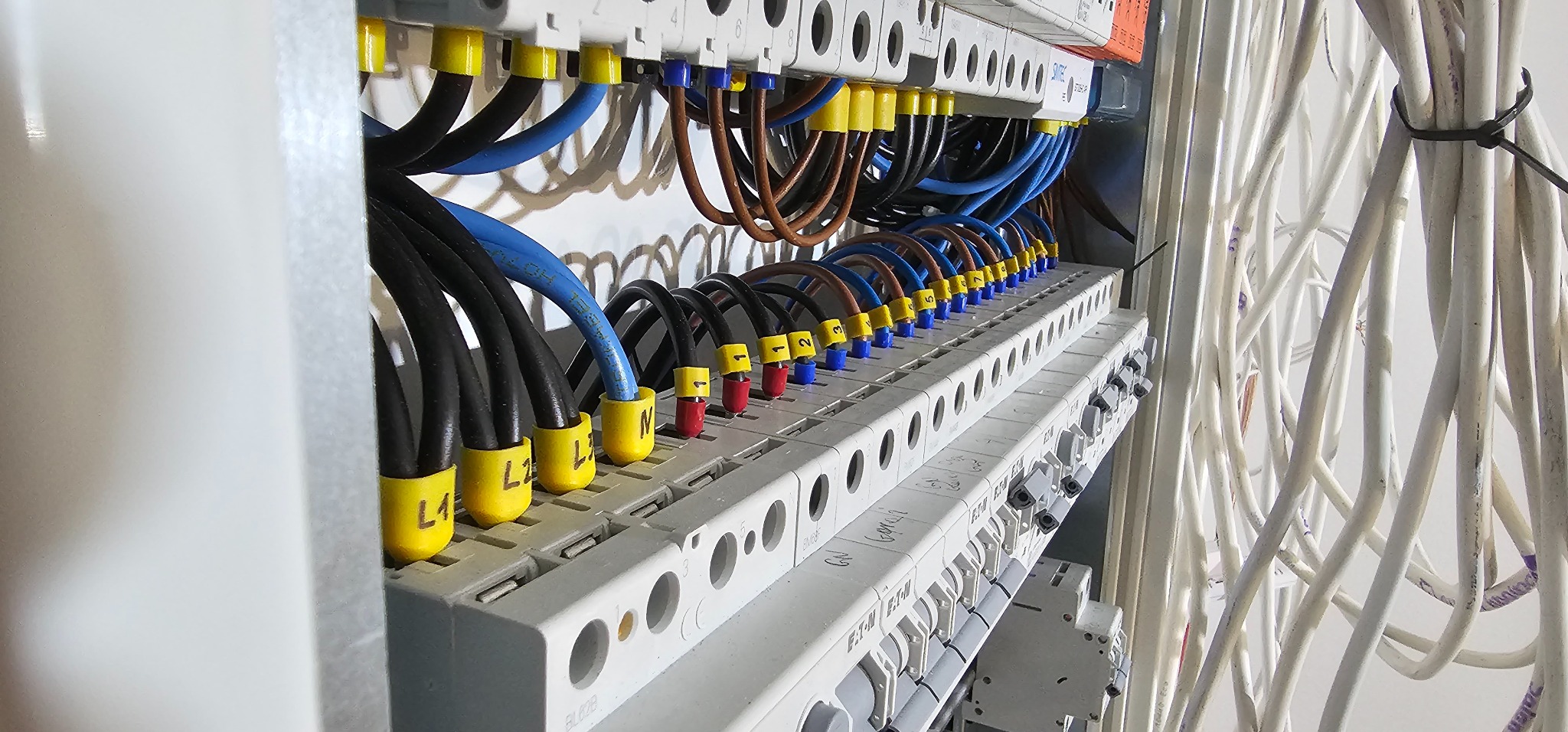Szczegółowe ujęcie tablicy rozdzielczej z podłączonymi przewodami oznaczonymi kolorowymi końcówkami i opisami L1, L2, L3, N; widoczne białe kable z boku, uporządkowane za pomocą opaski zaciskowej.