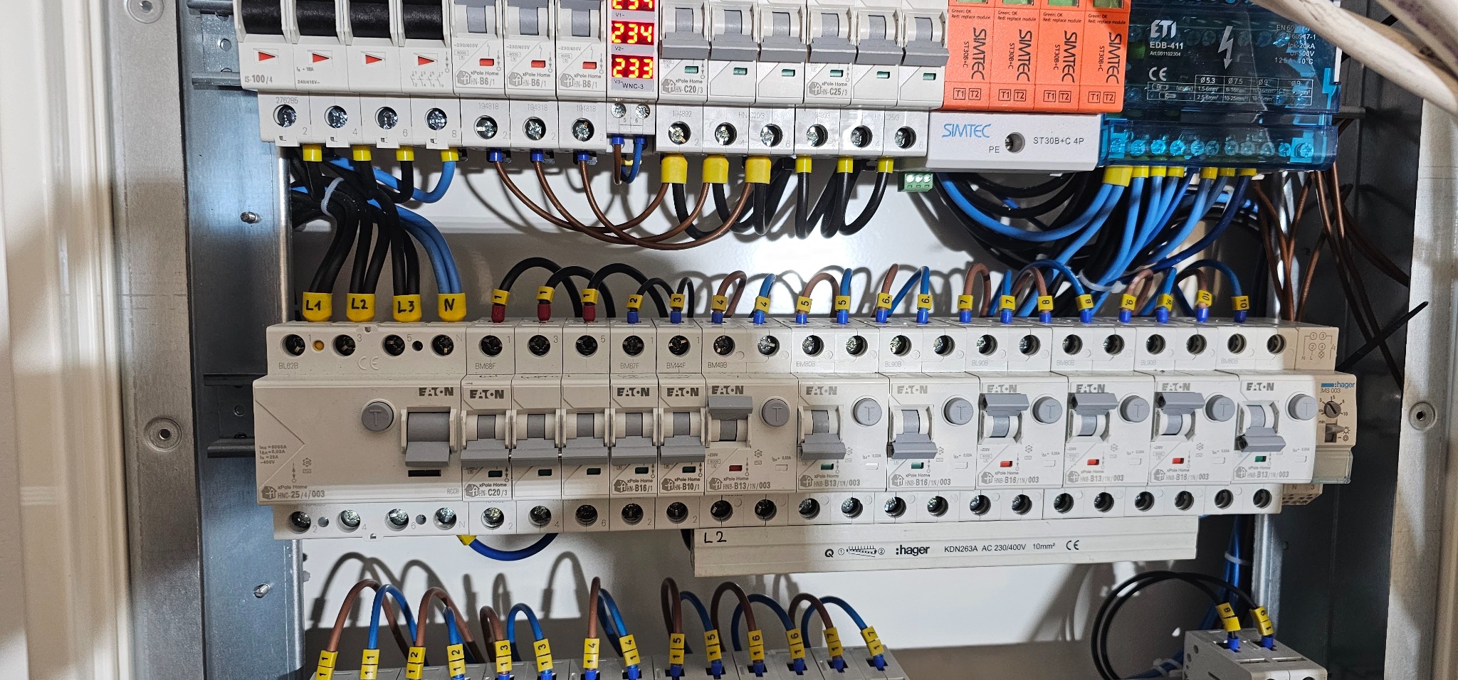Rozdzielnica elektryczna z widocznymi bezpiecznikami EATON, modułami SIMTEC, przewodami oznaczonymi numerami i wyświetlaczami cyfrowymi pokazującymi napięcie.