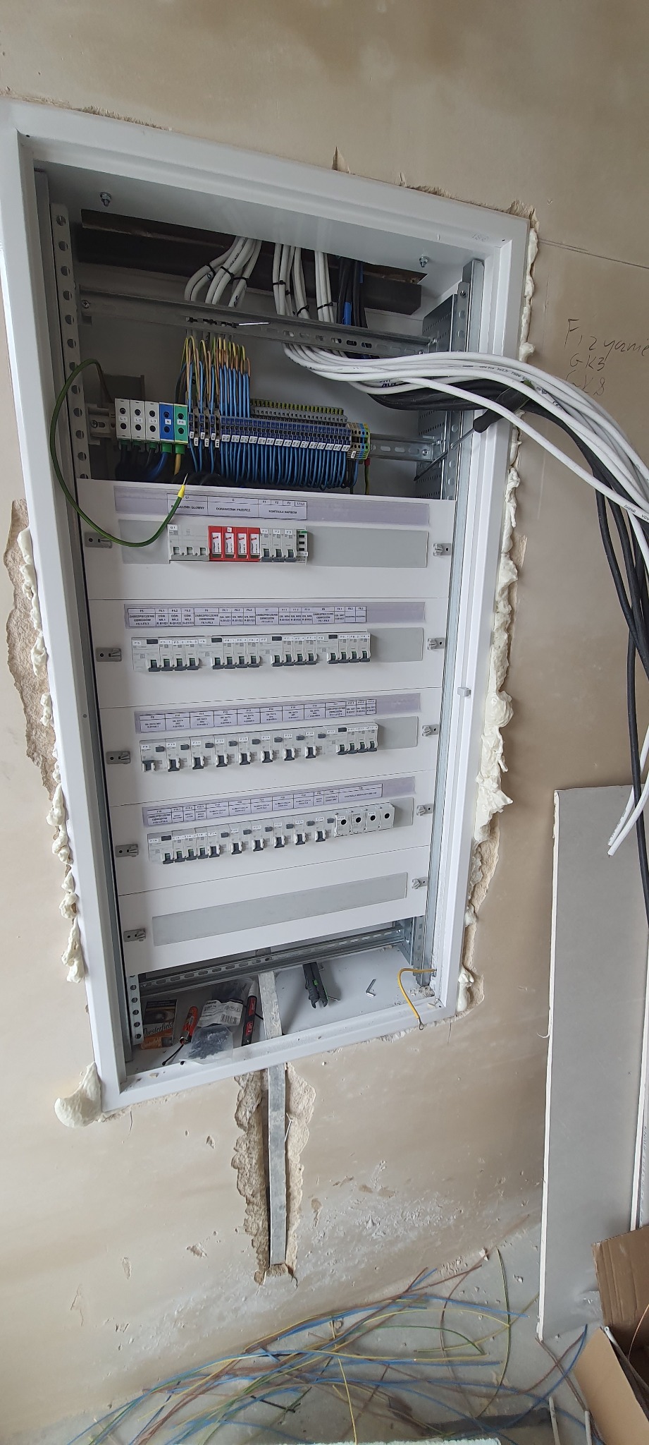 Rozdzielnica elektryczna z zamontowanymi bezpiecznikami i przewodami w trakcie instalacji, widoczne narzędzia i materiały montażowe, ściana w stanie surowym