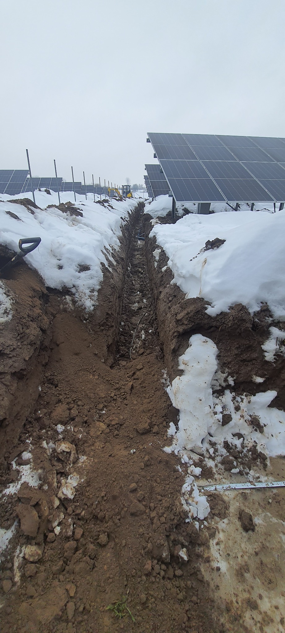 Wykop w ziemi częściowo pokrytej śniegiem, w tle panele słoneczne i mała koparka.