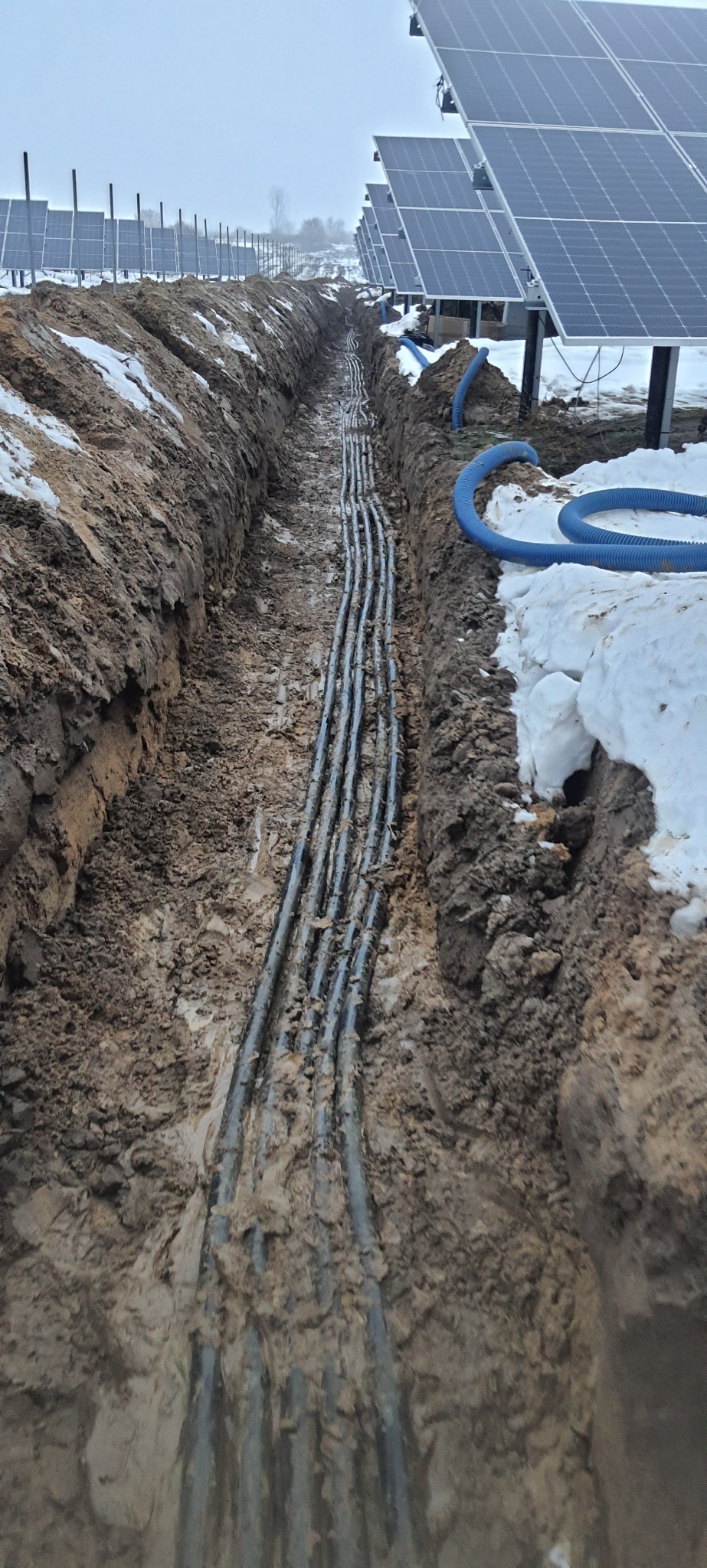 Wykop z ułożonymi kablami w ziemi, prawdopodobnie w trakcie realizacji instalacji fotowoltaicznej w Przyszowie, widoczne panele słoneczne w tle i fragment niebieskiego przewodu karbowanego.