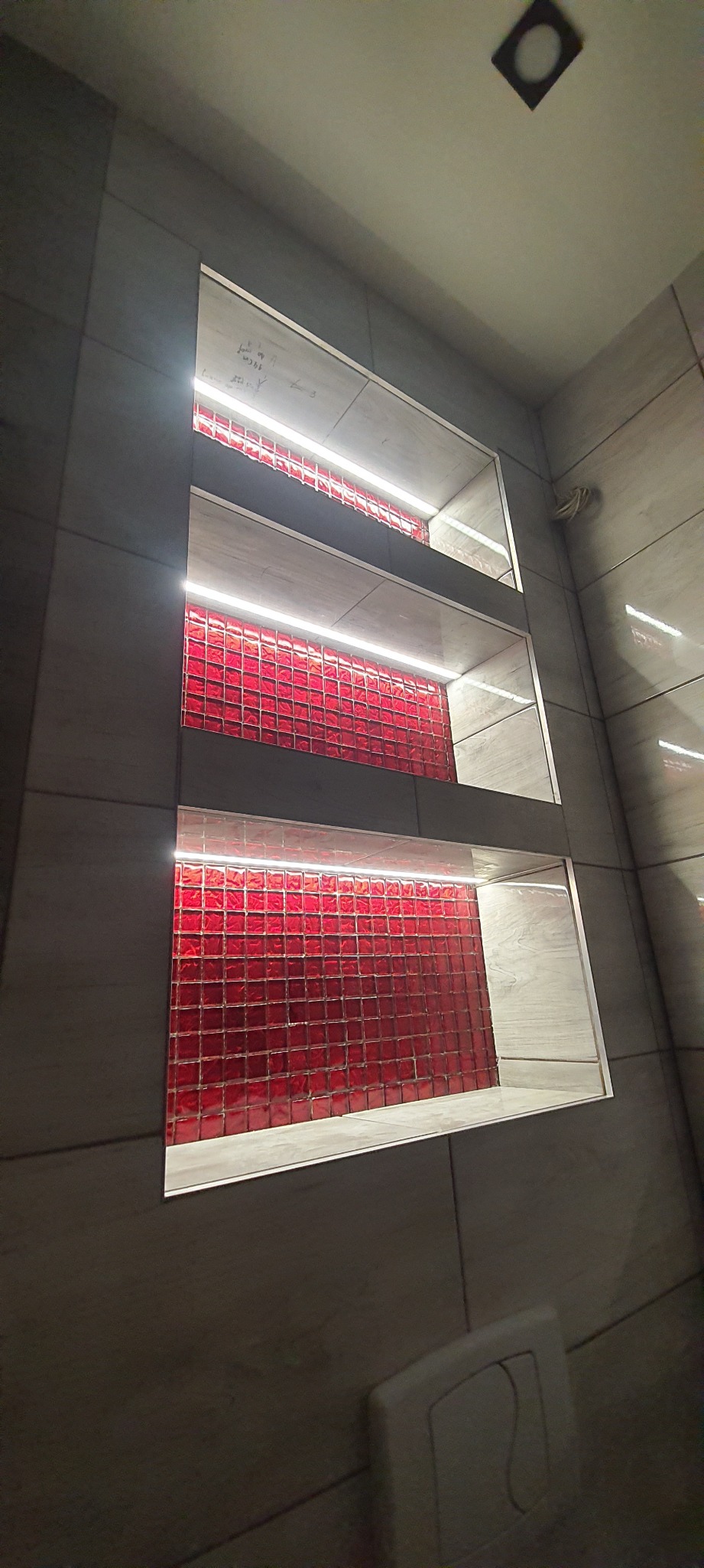 Narożne ujęcie ściany łazienki z trzema wnękami wyłożonymi czerwoną mozaiką i oświetleniem LED, widoczny fragment spłuczki toaletowej