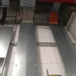 Kable elektryczne prowadzone w korytach metalowych pod sufitem z białych bloczków, widoczny czerwony rurociąg i uziemienie.