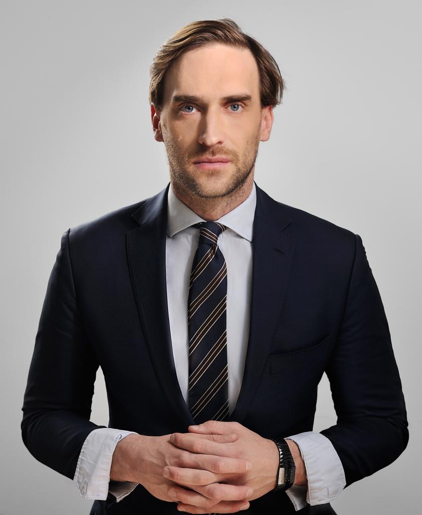 Portret mężczyzny w garniturze z krawatem w paski, splecione dłonie, zegarek na nadgarstku, na jasnym tle, neutralny wyraz twarzy, profesjonalny wizerunek.