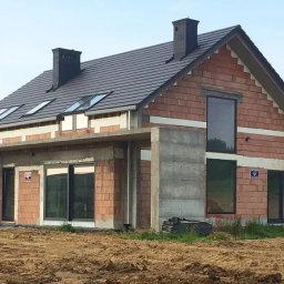N.i.s Paweł Jonatowski - Niedokończony dom jednorodzinny z czerwonej cegły z szarą dachówką, widoczne okna dachowe i betonowe elementy konstrukcyjne, teren wokół w stanie przygotowania.