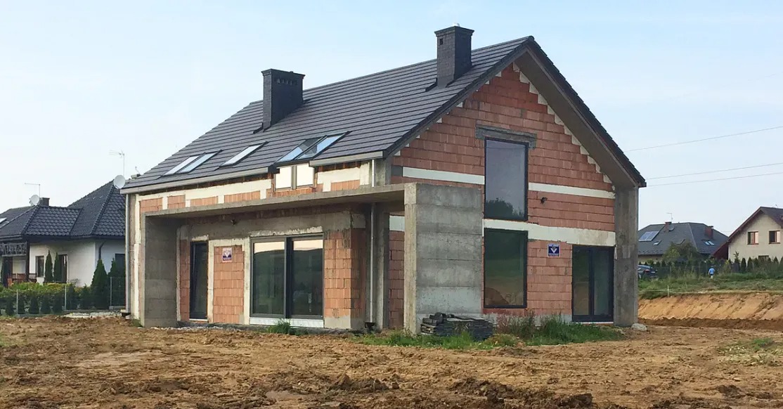 Niedokończony dom jednorodzinny z czerwonej cegły z szarą dachówką, widoczne okna dachowe i betonowe elementy konstrukcyjne, teren wokół w stanie przygotowania.