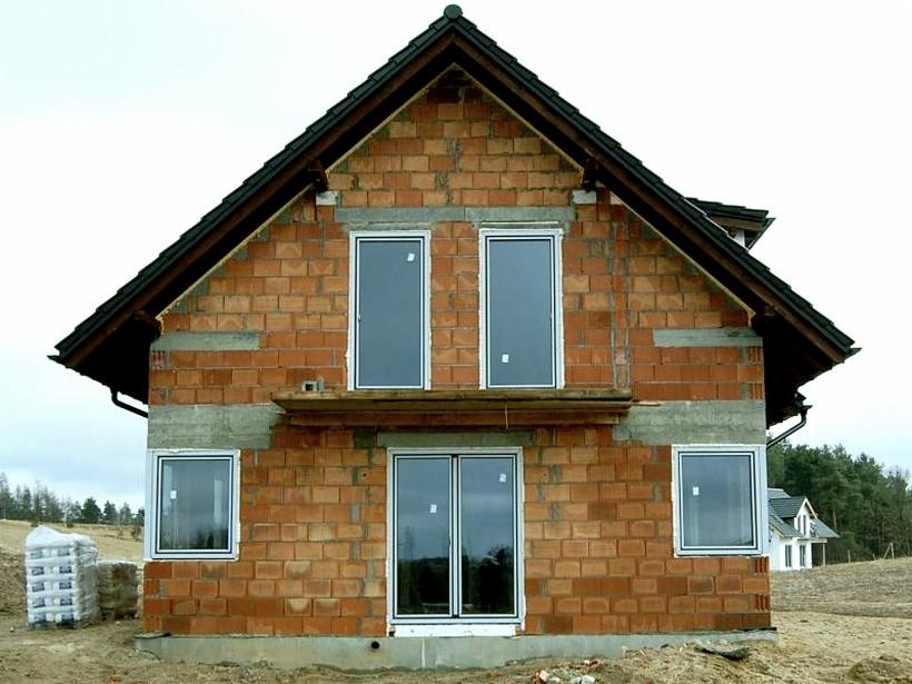 Surowy stan domu murowanego z widocznymi cegłami, zamontowanymi oknami z białymi ramami i stosem materiałów budowlanych obok.