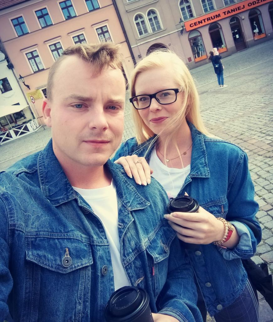 Para młodych ludzi w dżinsowych kurtkach robi sobie selfie na tle kamienic na starówce, trzymając kubki z kawą na wynos.