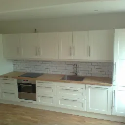 Jasna kuchnia z białymi szafkami, drewnianym blatem i białymi płytkami metra na ścianie, z widocznym zlewem i płytą indukcyjną.