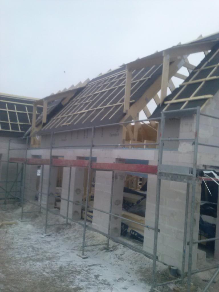 Budowa domu jednorodzinnego w stanie surowym otwartym: ściany z bloczków, drewniana konstrukcja dachu z pokryciem z papy, rusztowania wokół budynku.