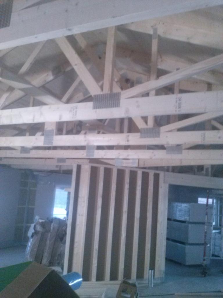 Wnętrze w trakcie budowy: widok na drewnianą konstrukcję dachu z metalowymi łącznikami, fragment ściany z pionowych desek i materiały budowlane w tle.