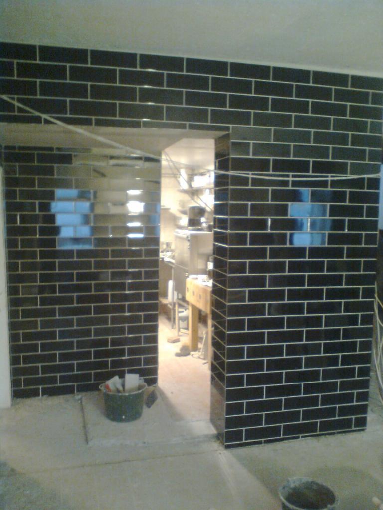 Częściowo wykończone obłożenie ścian czarną, błyszczącą glazurą w układzie cegiełek, widoczna przestrzeń wejściowa do pomieszczenia z wyposażeniem gastronomicznym w tle.