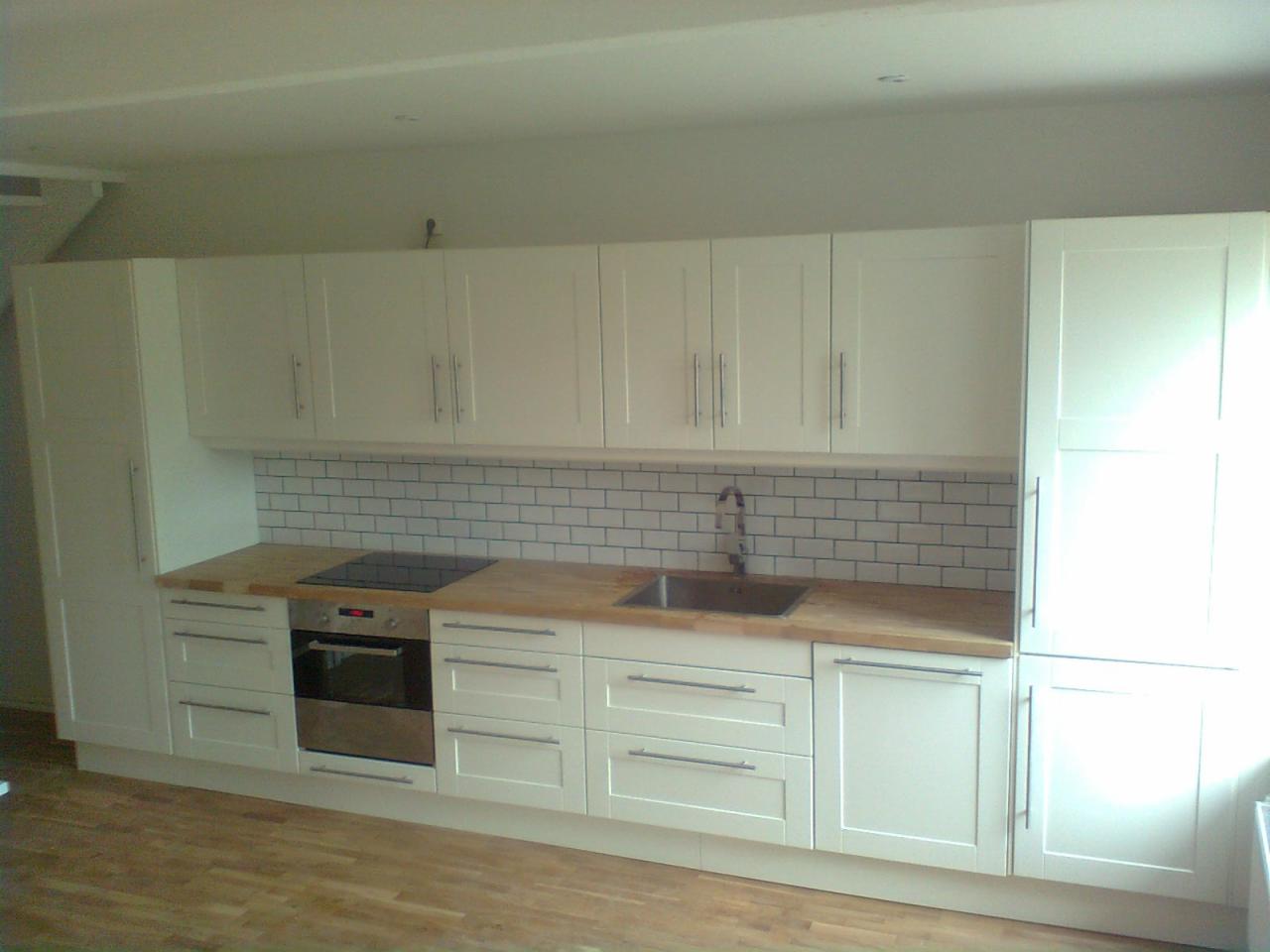 Jasna kuchnia z białymi szafkami, drewnianym blatem i białymi płytkami metra na ścianie, z widocznym zlewem i płytą indukcyjną.