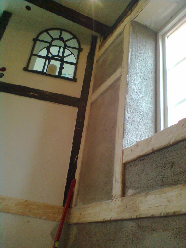 Wnętrze domu w trakcie prac wykończeniowych: surowe drewniane belki, okno w stylu retro i fragment ściany z nałożonym tynkiem strukturalnym, czerwona poziomica oparta o ścianę.