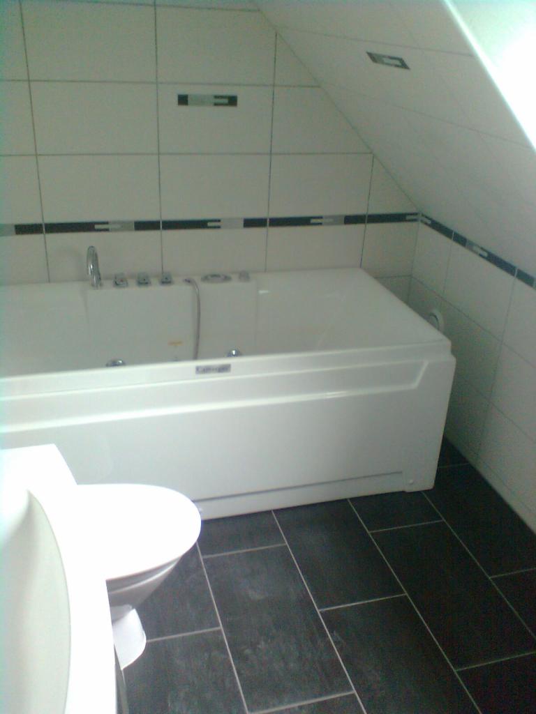 Biała wanna z hydromasażem w łazience z ukośnym sufitem, czarne płytki podłogowe i białe płytki ścienne z czarnym paskiem dekoracyjnym.