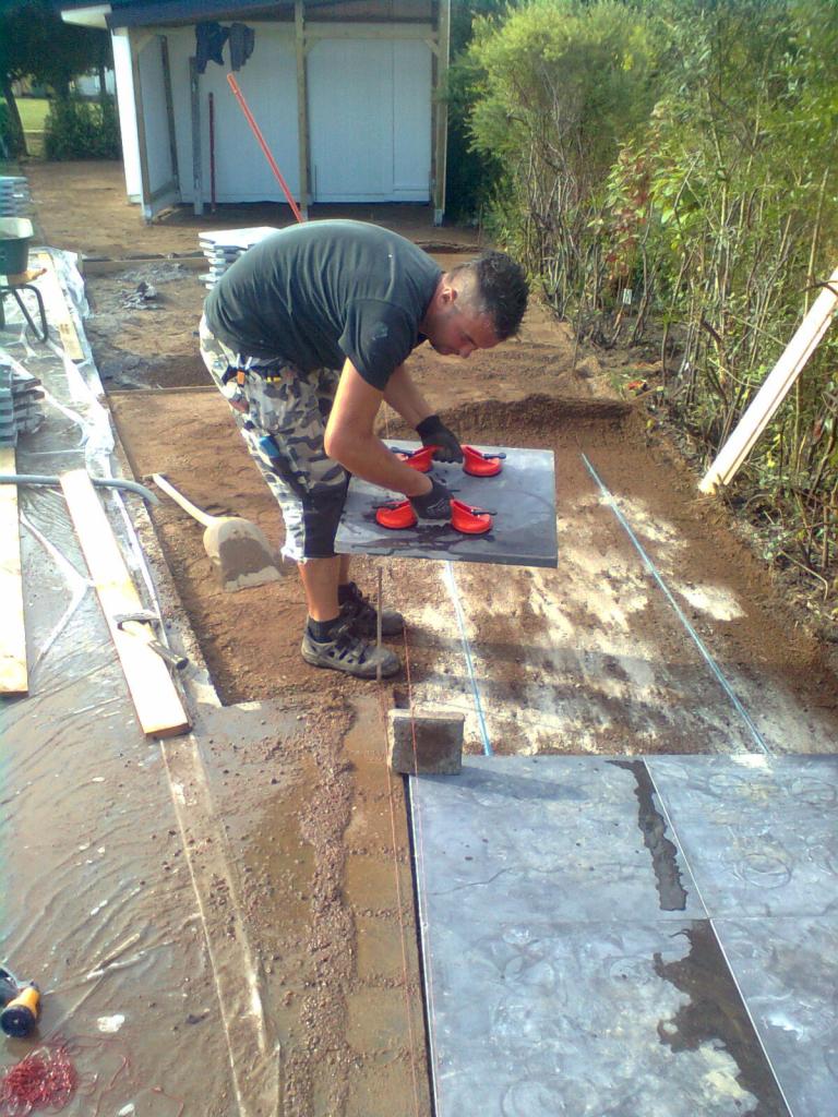 Pracownik w kamuflażowych spodniach układa dużą, szarą płytę chodnikową za pomocą przyssawek na przygotowanym podłożu, widoczne narzędzia i ustawione sznurki wyznaczające poziom.