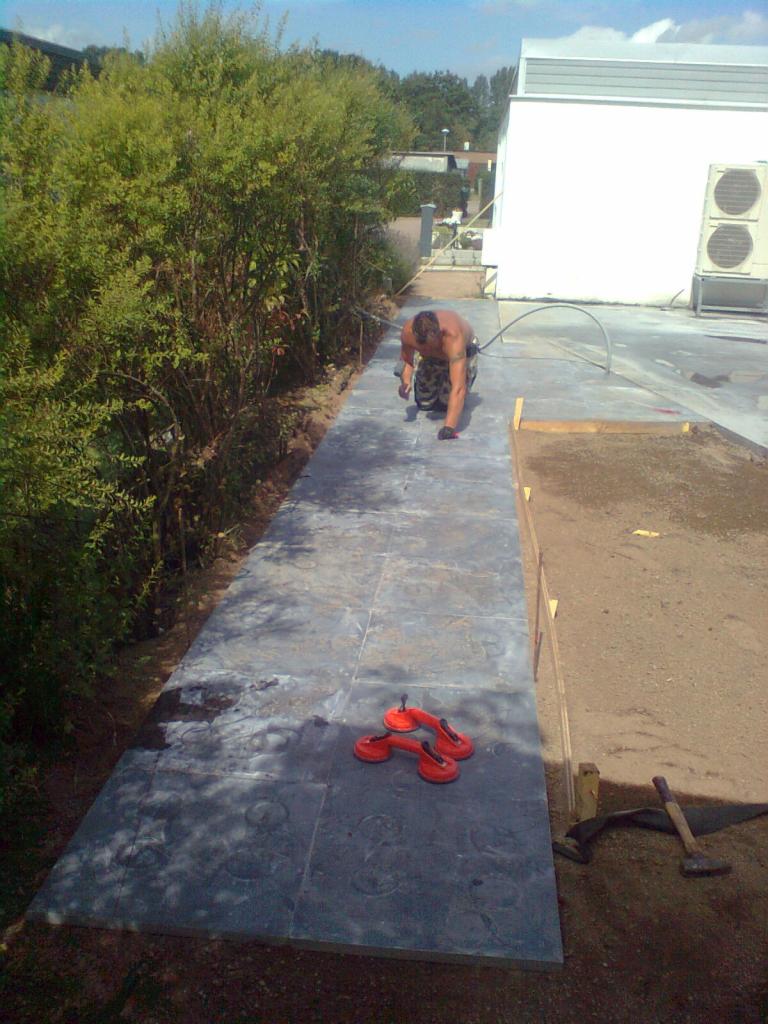 Mężczyzna w rękawicach układa ciemnoszare płyty chodnikowe na ścieżce obok żywopłotu, z użyciem przyssawki do przenoszenia płyt, w tle budynek z klimatyzacją.