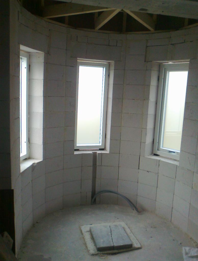 Wnętrze okrągłej konstrukcji w trakcie budowy z trzema oknami, widoczne białe bloczki, szara podłoga z płytą i fragmentem rury oraz drewniana konstrukcja dachu.