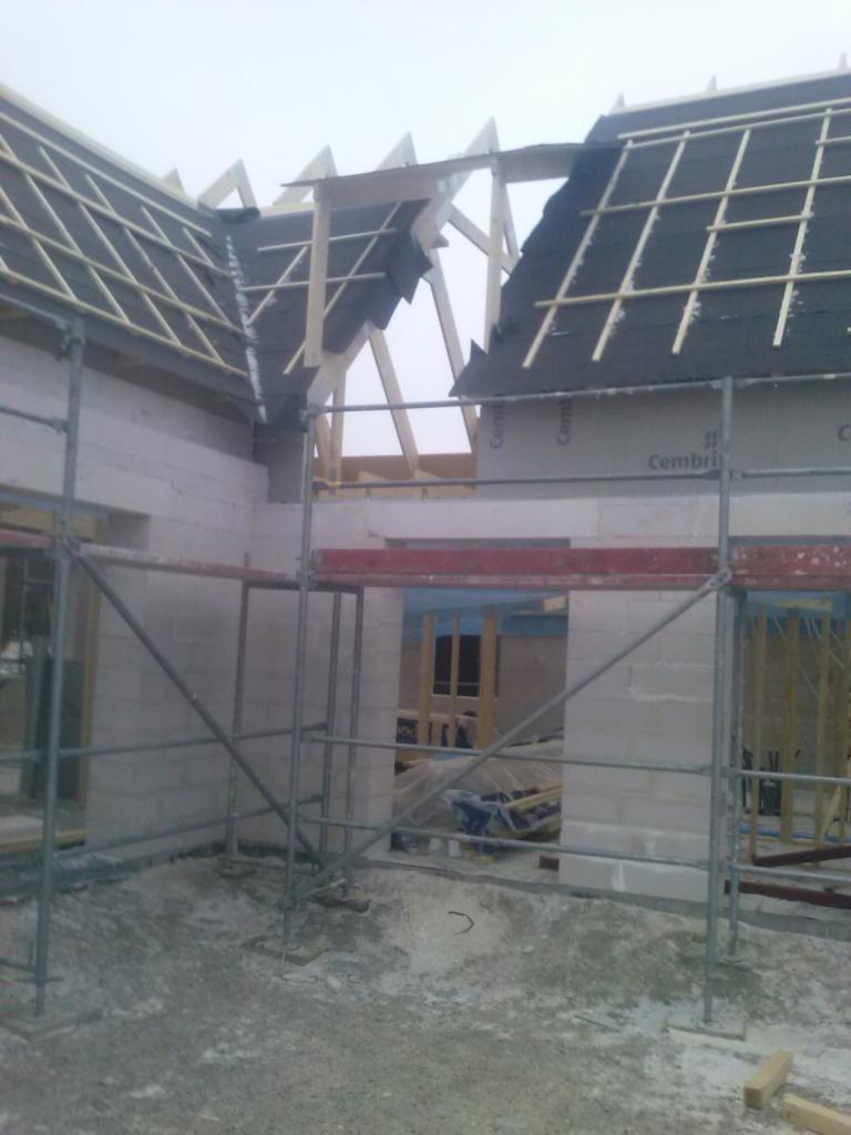 Budowa domu w trakcie wznoszenia dachu, widoczne rusztowania i bloczki betonowe, częściowo ułożona dachówka bitumiczna.