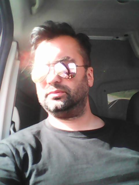 Mężczyzna w okularach przeciwsłonecznych siedzi w samochodzie, prawdopodobnie w trakcie jazdy, ubrany w czarną koszulkę. Widoczne odbicie w szkle.