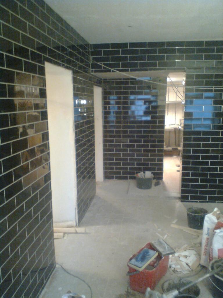 Wnętrze w trakcie wykańczania, ściany wyłożone błyszczącymi, czarnymi płytkami w układzie cegiełki, widoczne narzędzia i materiały budowlane, surowe wykończenie podłogi.