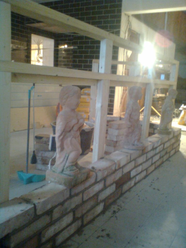 Wznoszony murek z cegły z trzema statuetkami aniołków, konstrukcja drewniana w tle, widoczne worki z zaprawą i narzędzia.