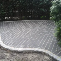 Ułożona nawierzchnia z ciemnoszarej kostki brukowej o nieregularnym kształcie, obrzeżona jasnoszarym kamieniem, otoczona zielenią.