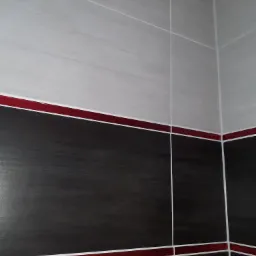 Wykończony narożnik łazienki z dwukolorowymi płytkami: górna część szara, dolna czarno-szara, oddzielone poziomym pasem z bordowych płytek.