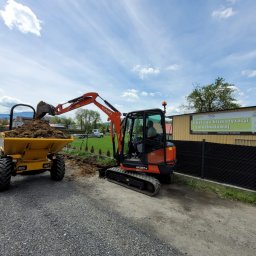 Pałka - Pomarańczowa minikoparka Kubota KX027-4 załadowuje ziemię do żółtej wywrotki na żwirowej drodze, widoczna reklama serwisu klimatyzacji samochodowej w tle.