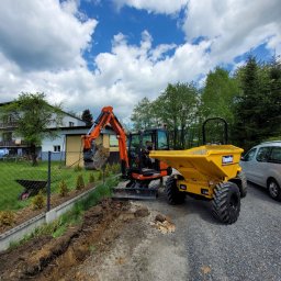 Pałka - Pomarańczowa mini koparka Kubota KX027-4 i żółta wywrotka Thwaites podczas prac ziemnych przy ogrodzeniu posesji, widoczny wykop i fragmenty cegieł.