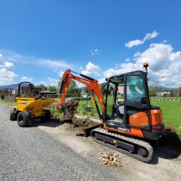 Pałka - Pomarańczowa mini koparka Kubota KX027-4 nabiera ziemię do żółtej wywrotki na tle wiejskiego krajobrazu z ogrodzeniem z siatki i domami.