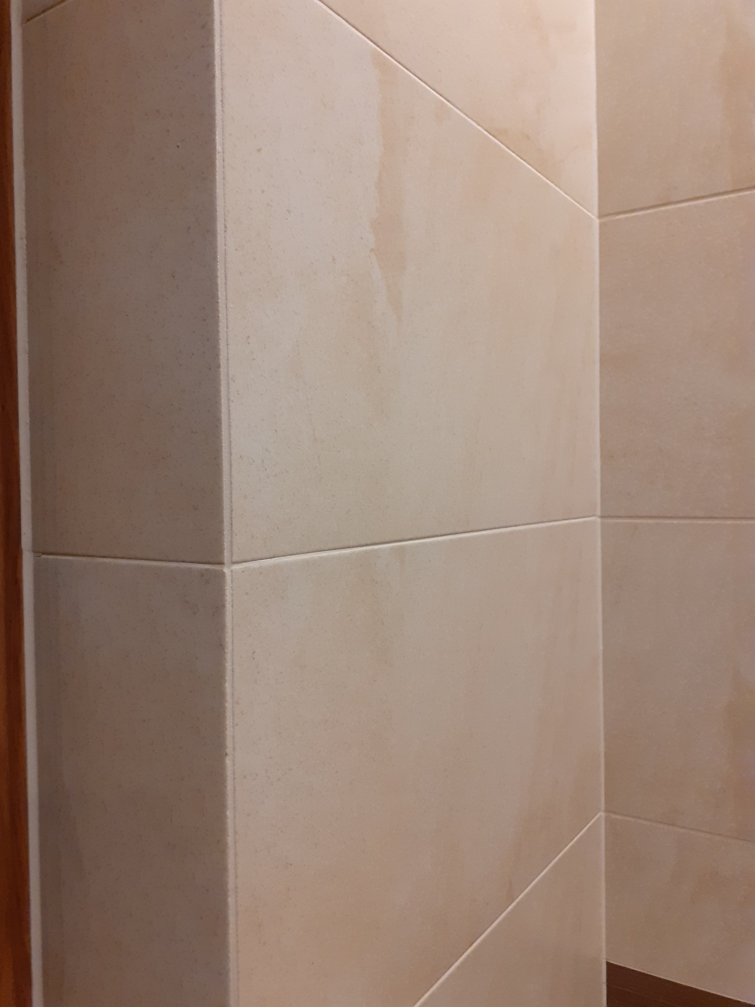 Fragment wykończonej ściany w łazience z beżowymi, kwadratowymi płytkami ceramicznymi, widoczne fugi i narożnik ściany.