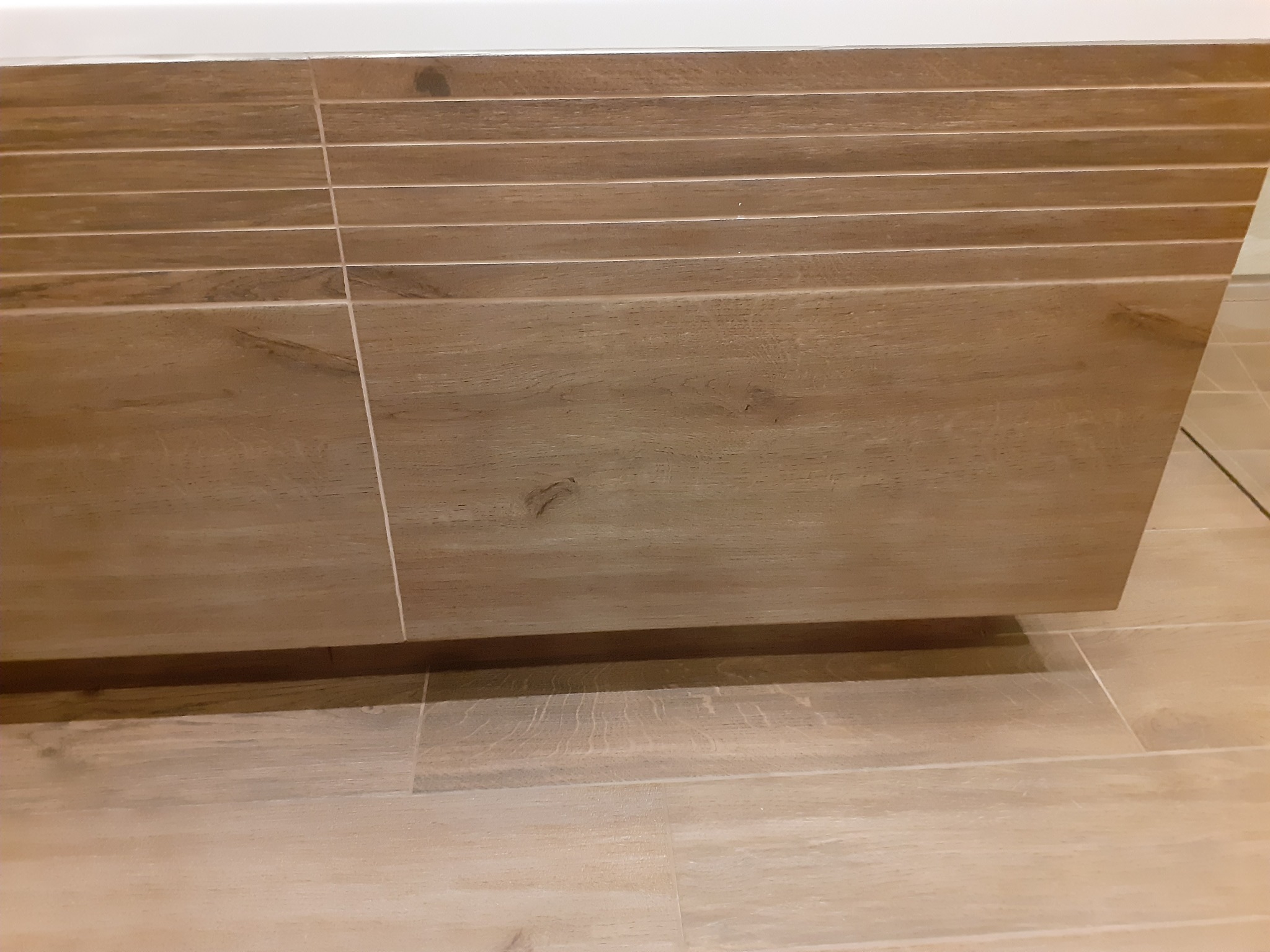 Szczegółowe ujęcie szafki łazienkowej z frontami imitującymi drewno, dopasowanymi kolorystycznie do płytek podłogowych, z widocznymi fugami. Szafka zawieszona, z delikatnym cieniem na podłodze.