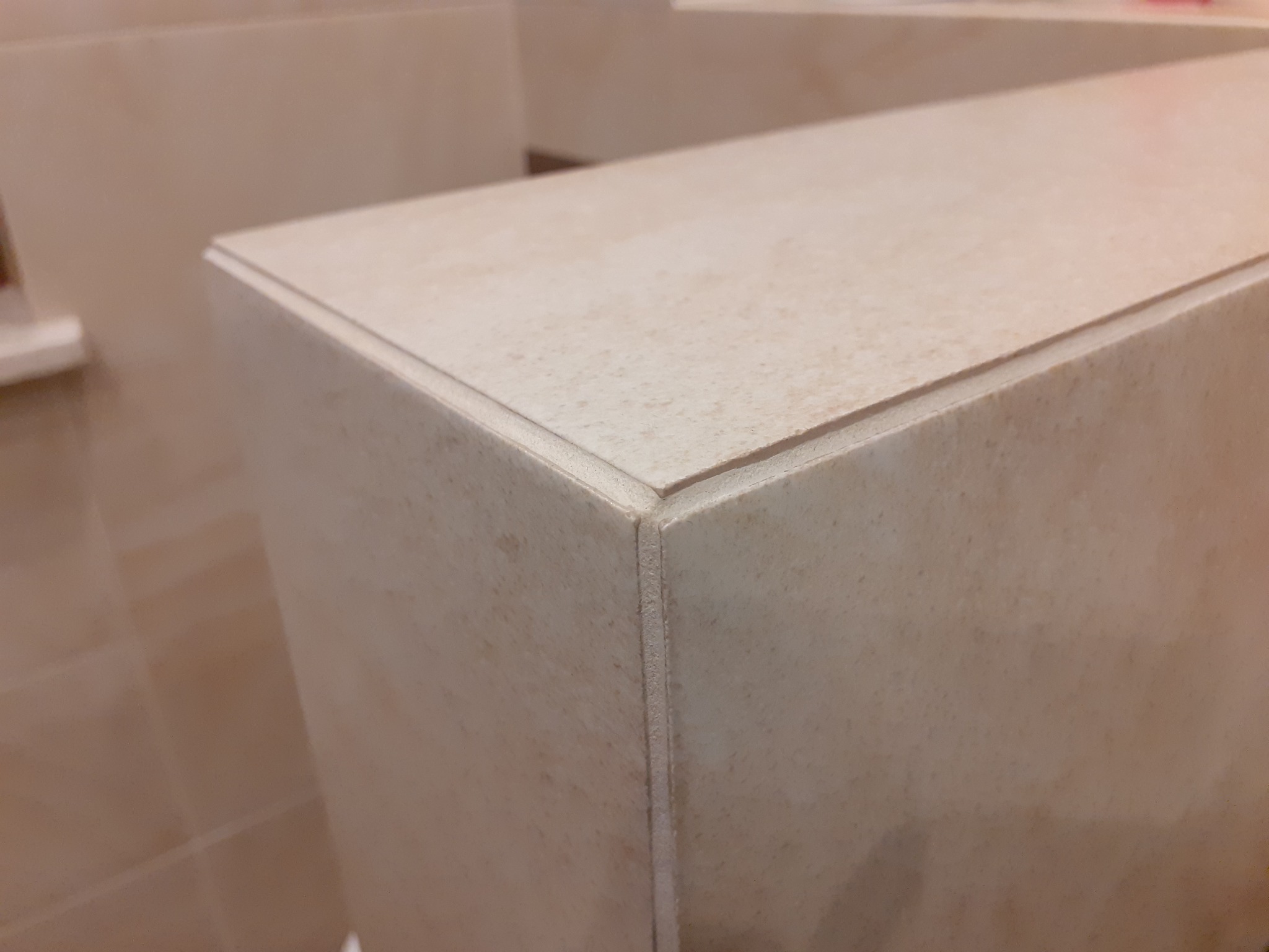 Detal narożnika obudowy w jasnych, matowych płytkach ceramicznych z widoczną fugą, prawdopodobnie w łazience.