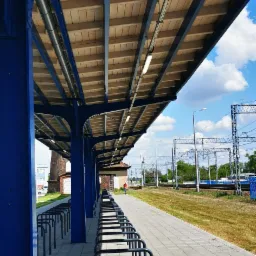 Zadaszony peron kolejowy z rzędami stojaków na rowery, oświetlony lampami jarzeniowymi, z widoczną konstrukcją stalową pomalowaną na niebiesko i drewnianym sufitem. W tle widoczne tory kolejowe...