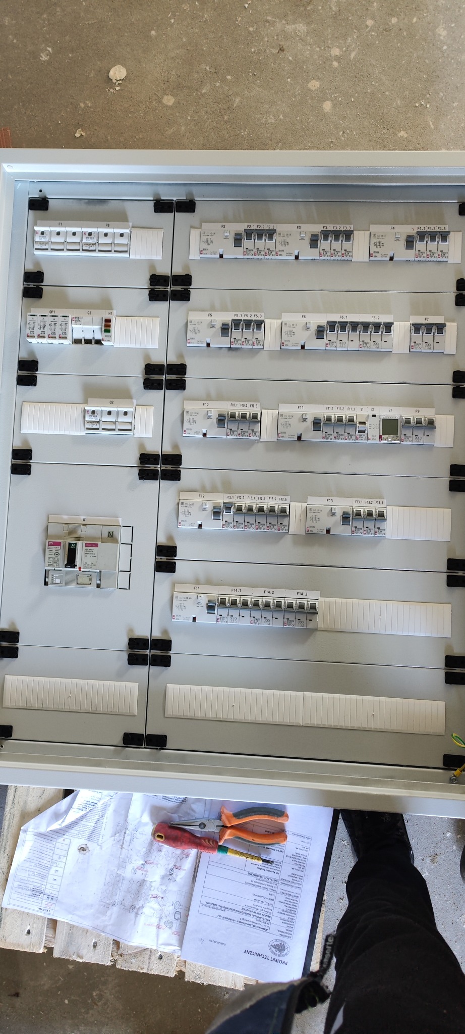 Rozdzielnica elektryczna z zainstalowanymi bezpiecznikami i wyłącznikami różnicowoprądowymi, widoczne narzędzia i dokumentacja projektowa na palecie pod spodem.