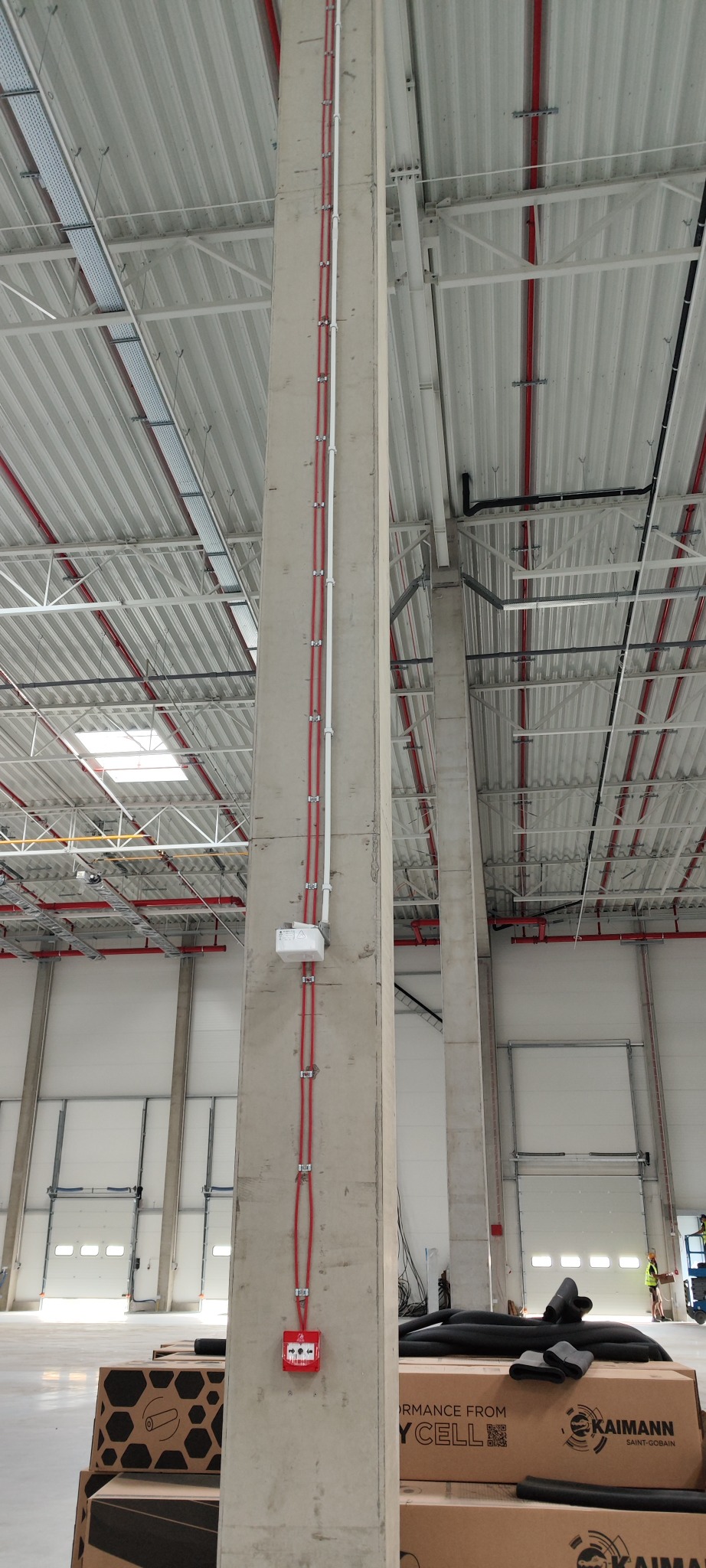 Betonowy słup w hali przemysłowej z poprowadzonymi pionowo przewodami elektrycznymi w czerwonej osłonie, białą rurą instalacyjną i zamontowanym na wysokości skrzynką oraz czerwonym przyciskiem...