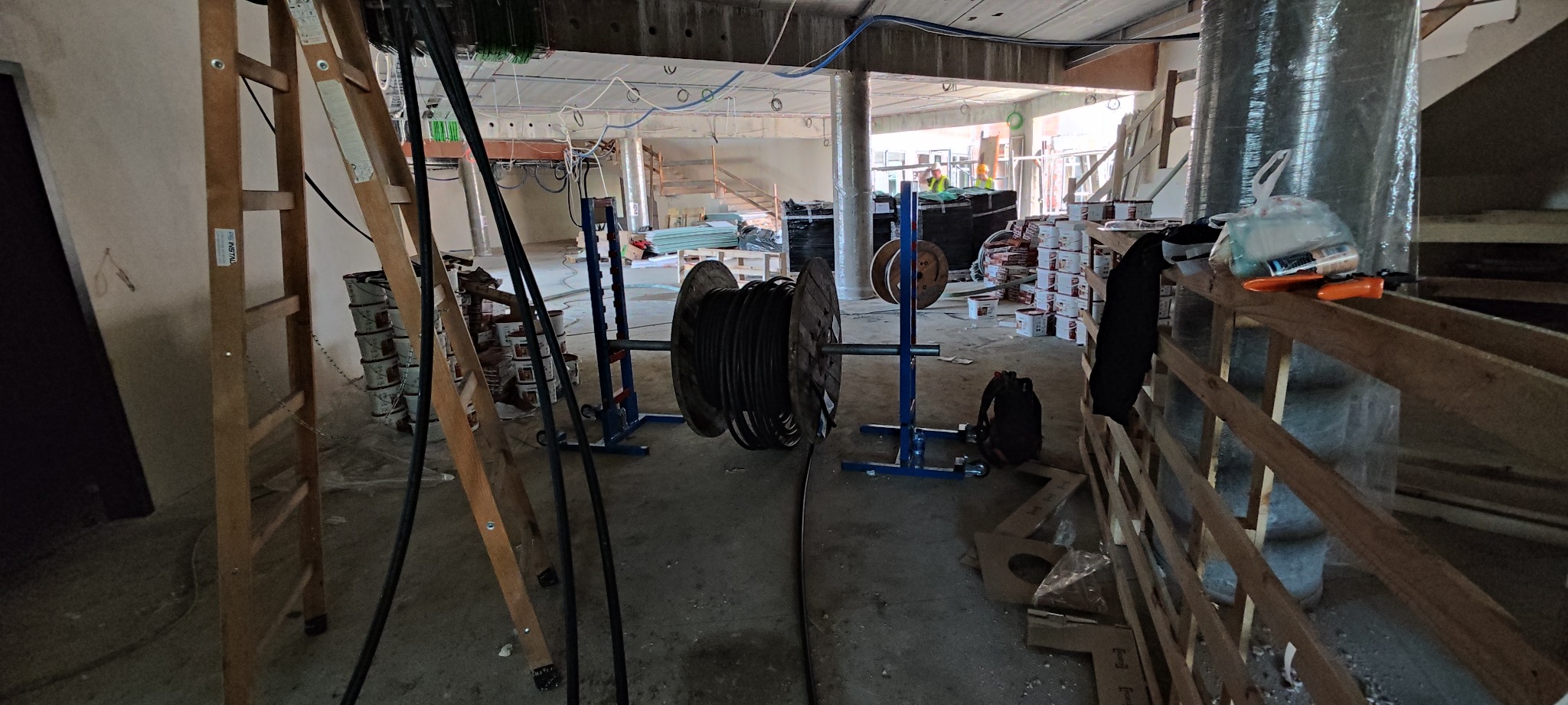 Wnętrze w trakcie instalacji elektrycznej: bębny z kablami na niebieskich stojakach, drabiny drewniane, puszki z farbą i widoczne przewody pod sufitem.