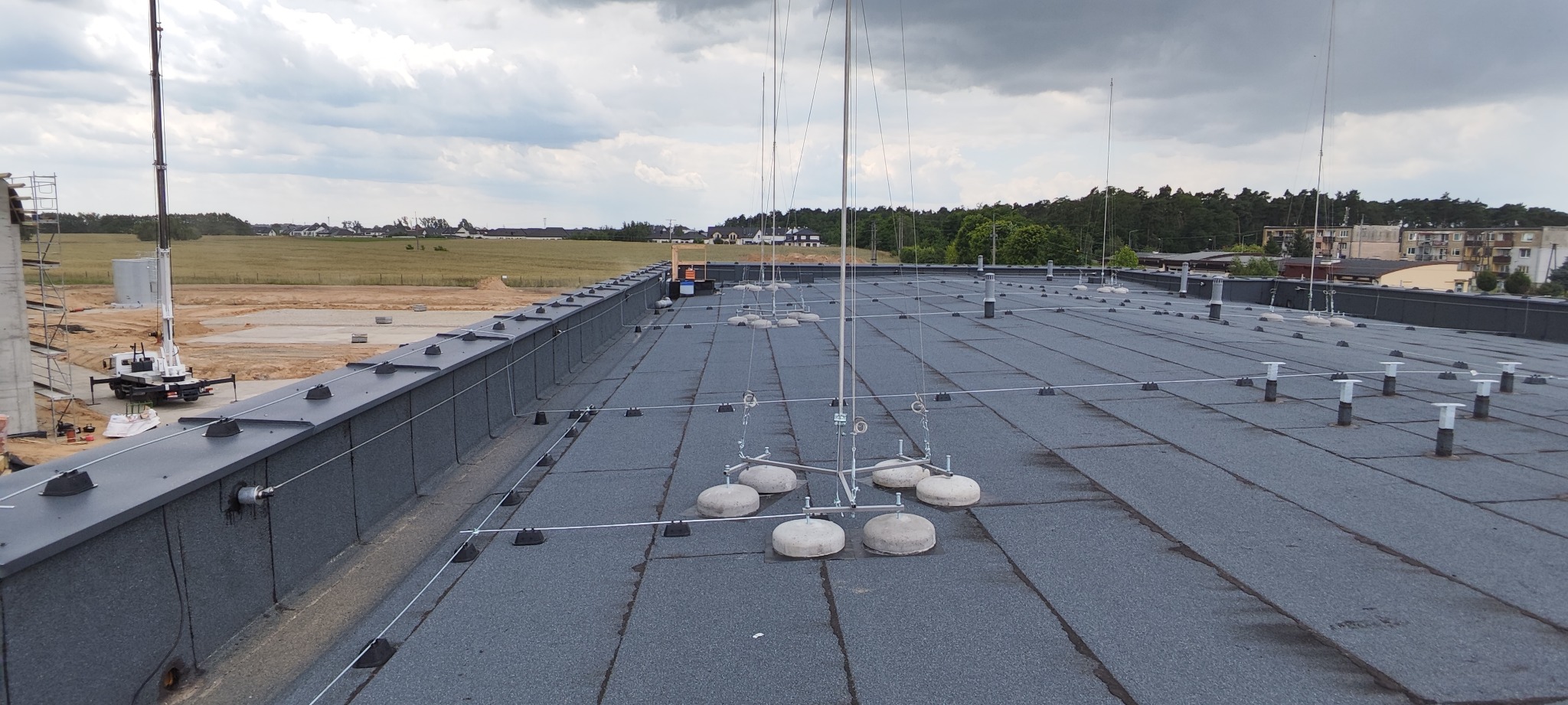 Rozległy dach przemysłowy z zainstalowaną siatką odgromową, widoczne betonowe podstawy masztów odgromowych i przewody, w tle budowa i krajobraz.