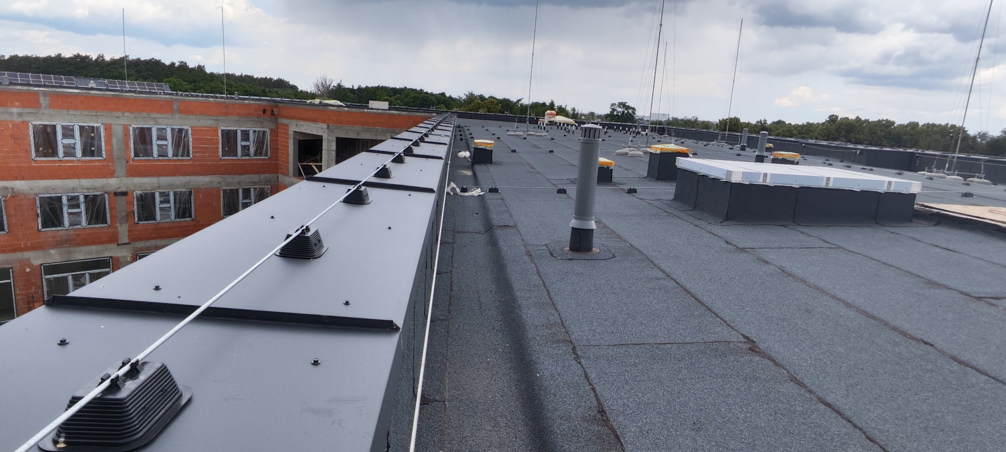 Rozległy dach pokryty papą z zainstalowanym systemem odgromowym, widoczne maszty odgromowe i przewody uziemiające, w tle budynek w trakcie budowy z widocznymi panelami fotowoltaicznymi na dachu.