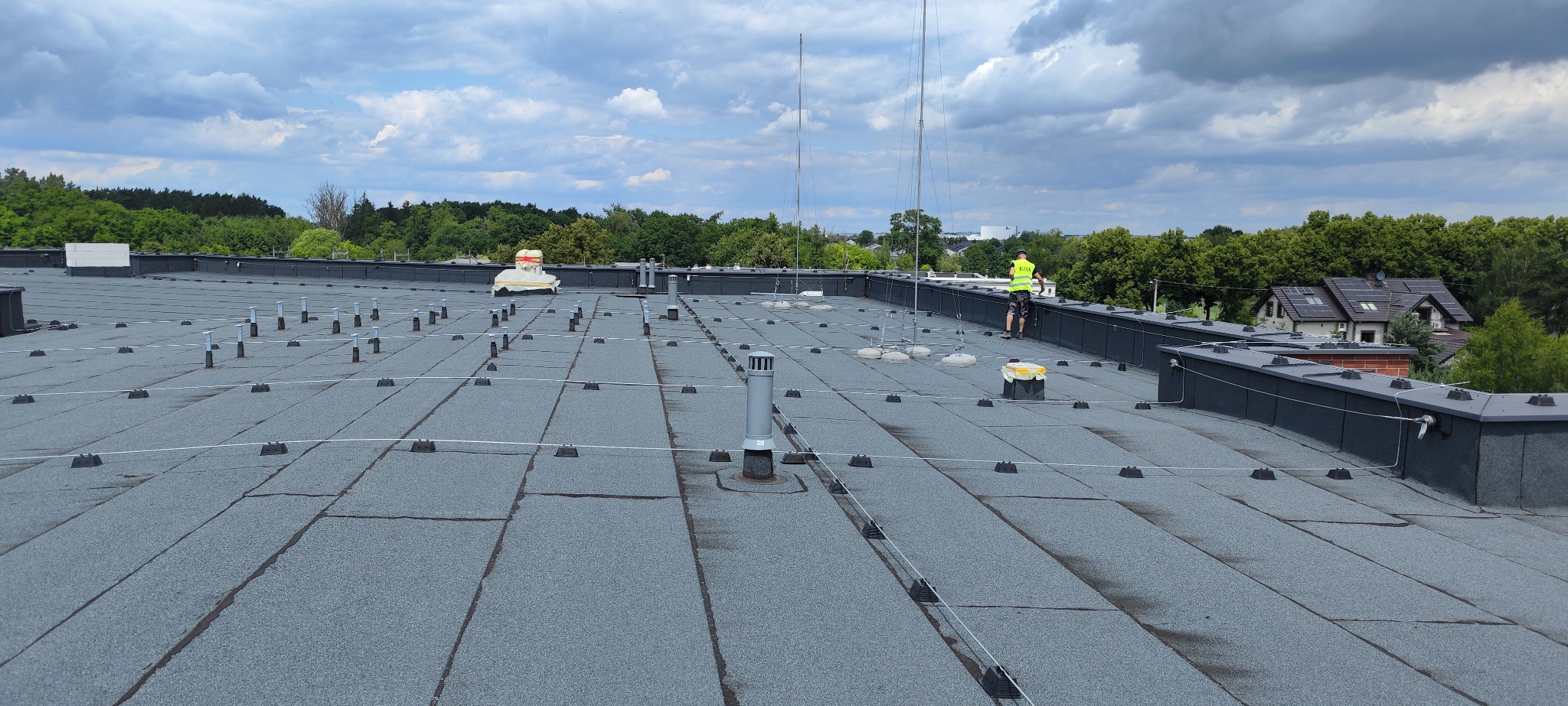 Rozległy dach pokryty papą z zamontowanymi uchwytami odgromowymi, widoczny pracownik w żółtej kamizelce przy krawędzi dachu oraz anteny odgromowe i kominy wentylacyjne.