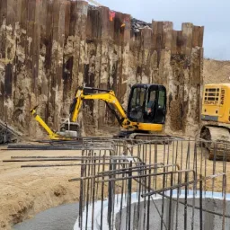 Budowa fundamentów: Dwa żółte koparki, w tym jedna marki Liebherr, pracują przy wykopie zabezpieczonym stalowymi ściankami Larsena. Widoczne zbrojenie fundamentu oraz materiały izolacyjne Izobud.