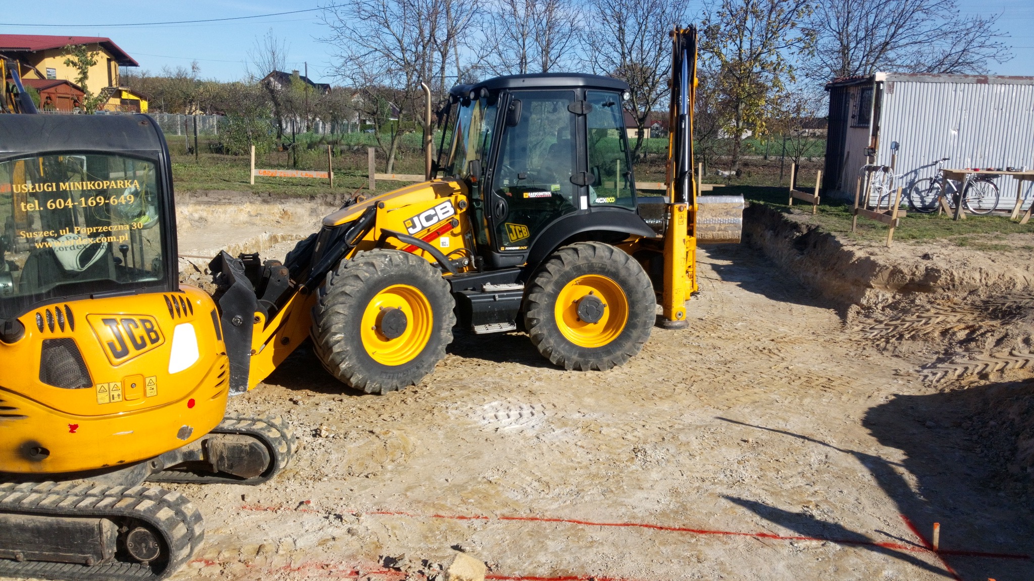 Żółta koparko-ładowarka JCB oraz minikoparka na placu budowy, widoczny wykop pod fundamenty z wytyczonym obrysem, w tle budynek gospodarczy i drzewa.