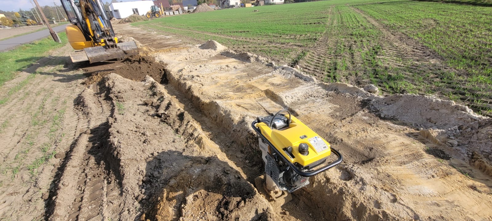Wykop pod instalacje z użyciem koparki i zagęszczarki, widoczne ślady opon na polu w tle.
