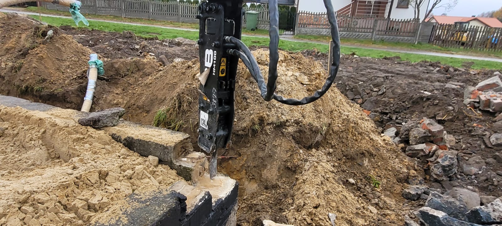 Hydrauliczny młot wyburzeniowy HM100 kruszy betonowy fundament w wykopie ziemnym; widoczne fragmenty starej konstrukcji i zasypany rurociąg.