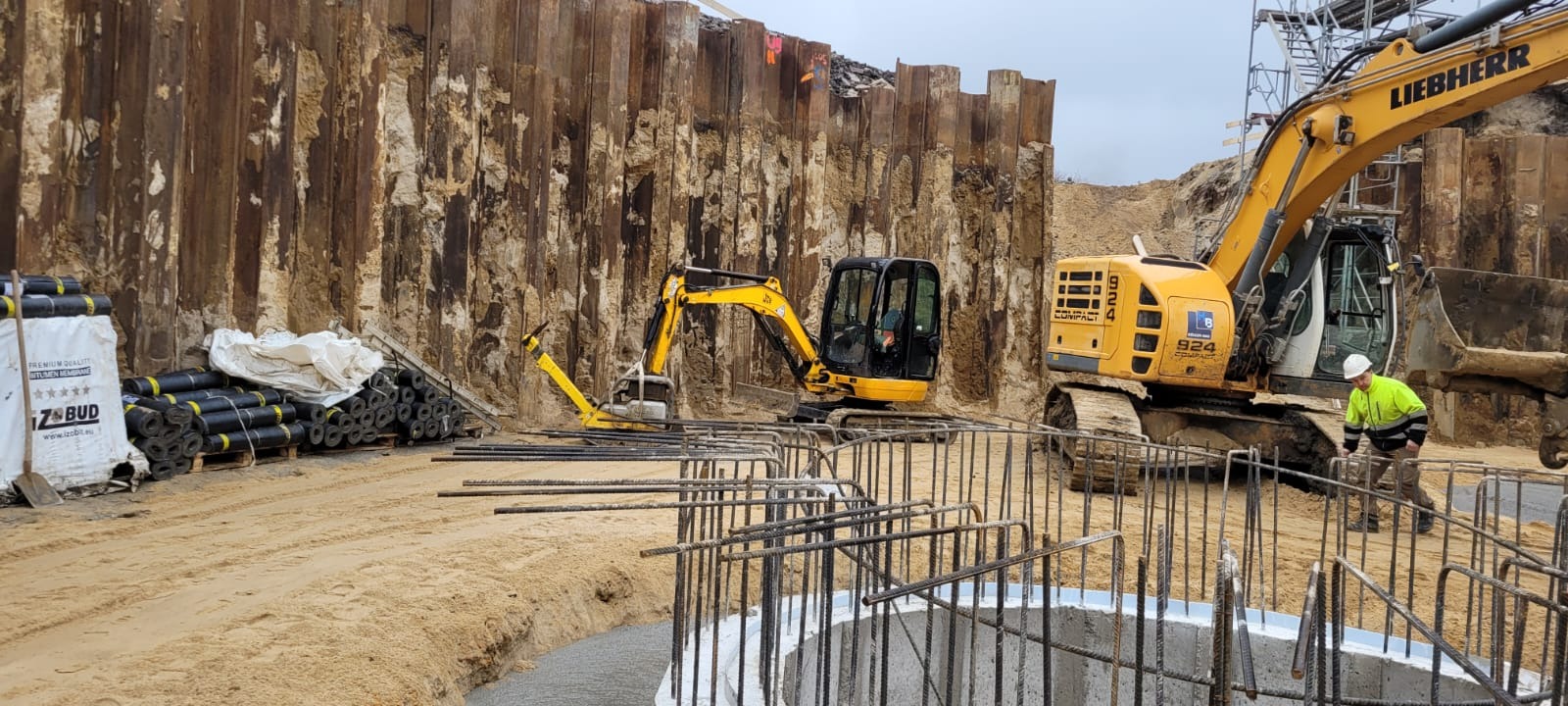 Budowa fundamentów: Dwa żółte koparki, w tym jedna marki Liebherr, pracują przy wykopie zabezpieczonym stalowymi ściankami Larsena. Widoczne zbrojenie fundamentu oraz materiały izolacyjne Izobud.