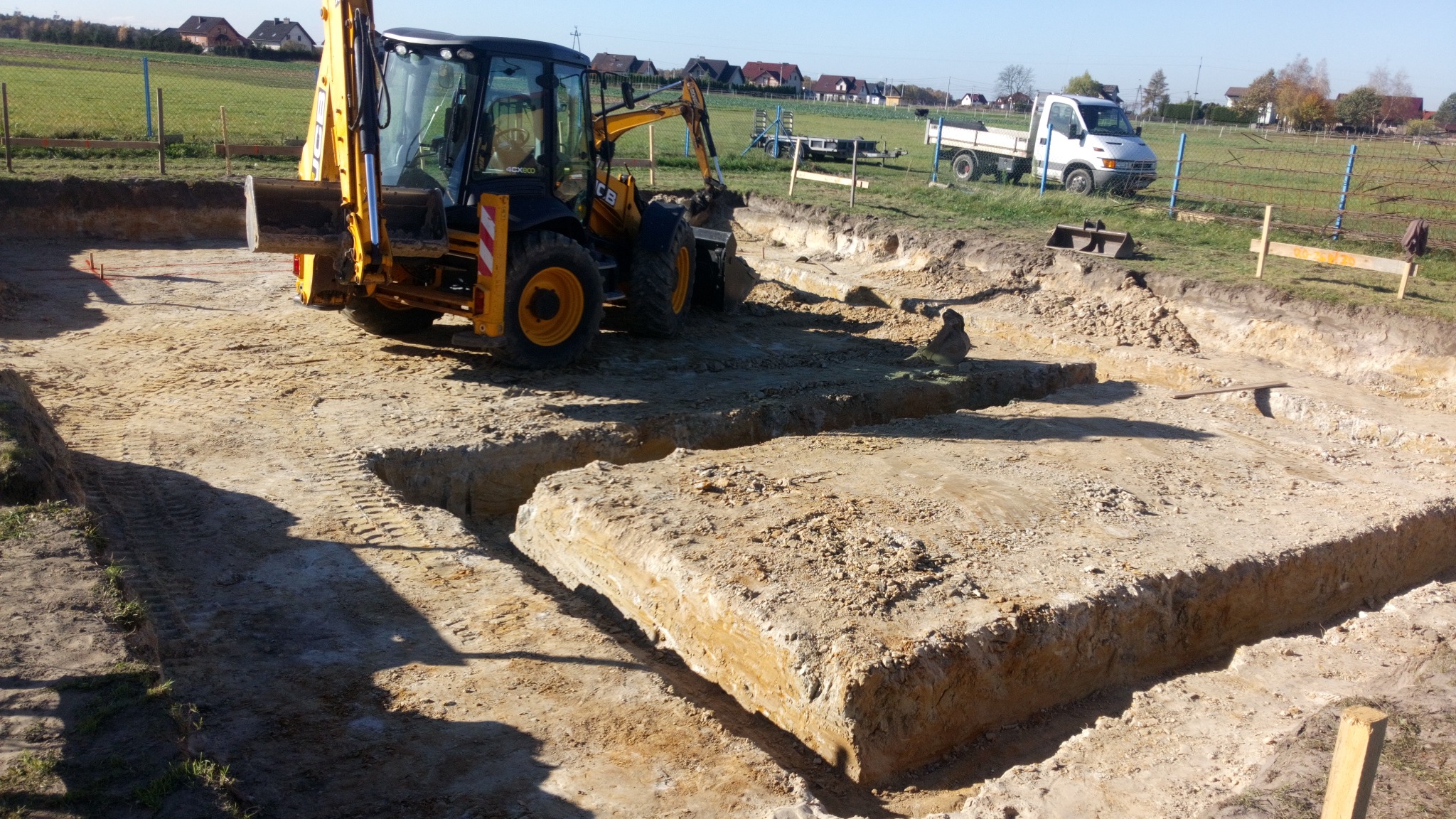 Żółta koparko-ładowarka JCB podczas prac ziemnych przy wykopie pod fundamenty; widoczne przygotowane zbrojenie i odkładana ziemia w tle, niebo bezchmurne.