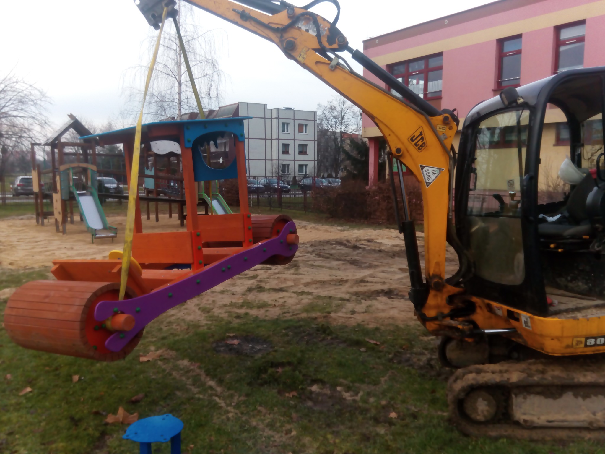 Żółta koparka JCB podnosi za pomocą pasów kolorowy, drewniany walec do zabawy dla dzieci na placu zabaw; w tle budynki mieszkalne i drzewa bez liści.