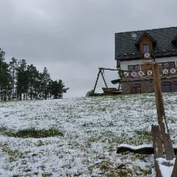 Zimowy krajobraz ośrodka wypoczynkowego z drewnianym domem, huśtawką i boiskiem do siatk&oacute;wki w tle, z pierwszym planem fragmentu konstrukcji drewnianej pokrytej śniegiem.